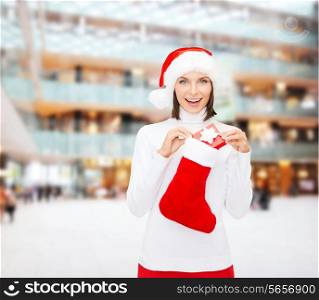 christmas, winter, happiness, holidays and people concept - smiling woman in santa helper hat with small gift box and stocking over shopping center background