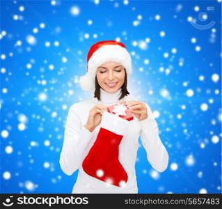 christmas, winter, happiness, holidays and people concept - smiling woman in santa helper hat with small gift box and stocking over blue snowy background