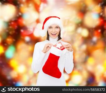 christmas, winter, happiness, holidays and people concept - smiling woman in santa helper hat with small gift box and stocking over red lights background