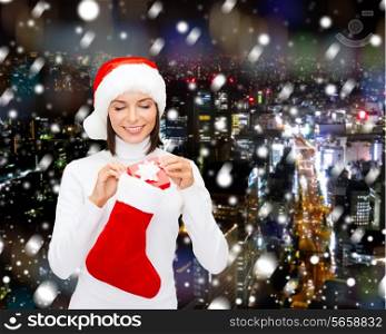christmas, winter, happiness, holidays and people concept - smiling woman in santa helper hat with small gift box and stocking over snowy night city background