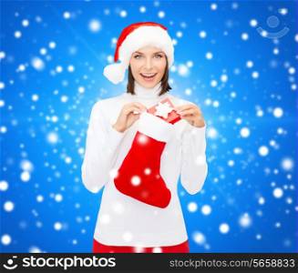 christmas, winter, happiness, holidays and people concept - smiling woman in santa helper hat with small gift box and stocking over blue snowy background