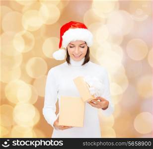 christmas, winter, happiness, holidays and people concept - smiling woman in santa helper hat opening gift box over beige lights background