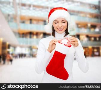 christmas, winter, happiness, holidays and people concept - smiling woman in santa helper hat with small gift box and stocking over shopping center background