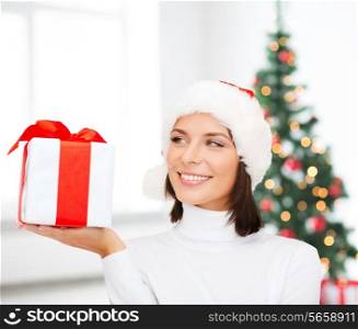 christmas, winter, happiness, holidays and people concept - smiling woman in santa helper hat with gift box over living room and christmas tree background