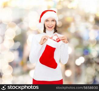 christmas, winter, happiness, holidays and people concept - smiling woman in santa helper hat with small gift box and stocking over lights background