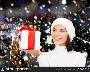 christmas, winter, happiness, holidays and people concept - smiling woman in santa helper hat with gift box over snowy night city background