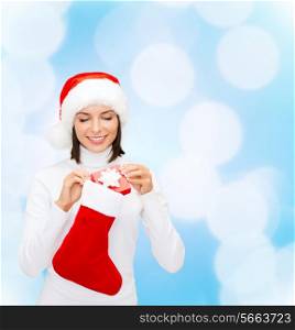 christmas, winter, happiness, holidays and people concept - smiling woman in santa helper hat with small gift box and stocking over blue lights background