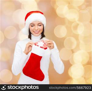 christmas, winter, happiness, holidays and people concept - smiling woman in santa helper hat with small gift box and stocking over beige lights background