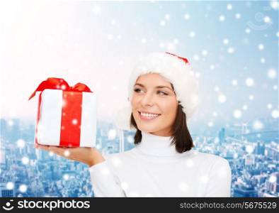 christmas, winter, happiness, holidays and people concept - smiling woman in santa helper hat with gift box over snowy city background