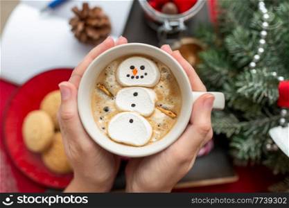 christmas, winter holidays and leisure concept - close up of hands holding cup of coffee with marshmallow snowman. hands with marshmallow snowman in mug on christmas