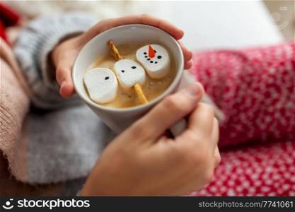 christmas, winter holidays and leisure concept - close up of hands holding cup of coffee with marshmallow snowman at home. hands with marshmallow snowman in mug on christmas