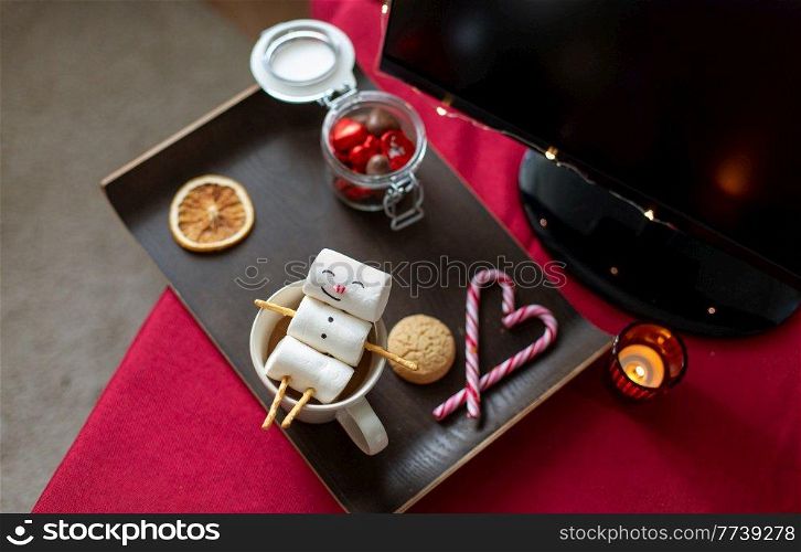 christmas, winter holidays and leisure concept - close up of marshmallow snowman taking bath in cup of coffee, sweet treats and tv monitor on table with red cloth at home. marshmallow snowman in cup of coffee on christmas