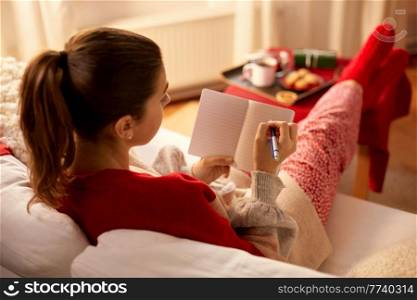 christmas, winter holidays and leisure concept - close up of young woman with pencil writing to notebook and resting her feet on table at cozy home. woman writing to notebook at home on christmas