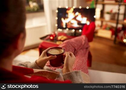 christmas, winter holidays and leisure concept - close up of young woman watching tv with fireplace on screen and drinking coffee with her feet on table at cozy home. woman watching tv and drinking coffee on christmas