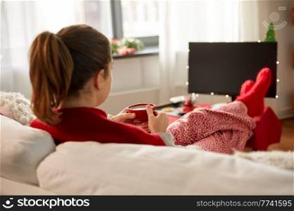 christmas, winter holidays and leisure concept - close up of young woman watching tv and drinking coffee with her feet on table at cozy home. woman watching tv and drinking coffee on christmas