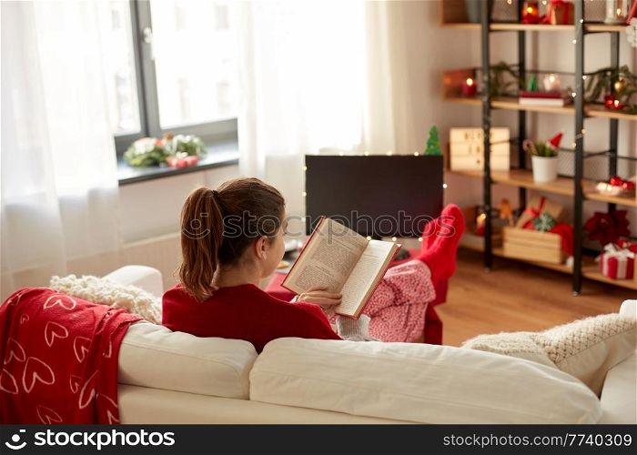 christmas, winter holidays and leisure concept - young woman reading book and resting her feet on table at cozy home. young woman reading book at home on christmas