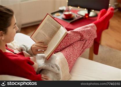 christmas, winter holidays and leisure concept - young woman reading book and resting her feet on table at cozy home. young woman reading book at home on christmas