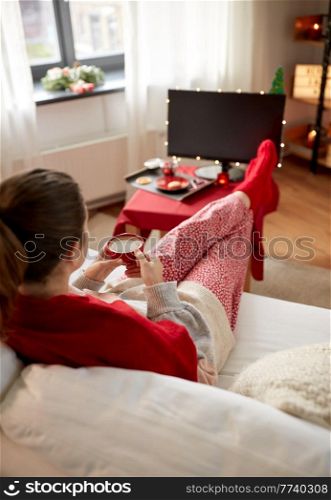 christmas, winter holidays and leisure concept - young woman watching tv and drinking coffee with her feet on table at cozy home. woman watching tv and drinking coffee on christmas