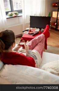 christmas, winter holidays and leisure concept - young woman watching tv and drinking coffee with her feet on table at cozy home. woman watching tv and drinking coffee on christmas