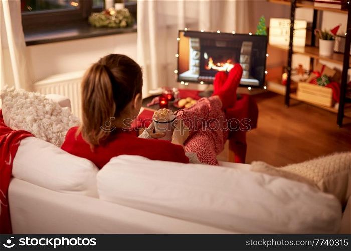 christmas, winter holidays and leisure concept - young woman watching tv with fireplace on screen and holding mug of marshmallow and whipped cream resting feet on table at cozy home. woman watching tv with cream and marshmallow on christmas