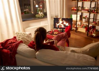 christmas, winter holidays and leisure concept - young woman watching tv with fireplace on screen and drinking coffee with her feet on table at cozy home. woman watching tv and drinking coffee on christmas