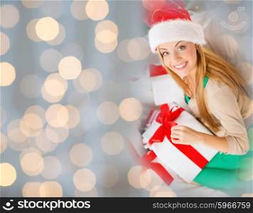 christmas, winter, holidays and people concept - happy woman in santa hat with christmas gifts over lights background