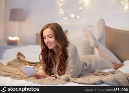 christmas, winter, holidays and people concept - happy young woman with pencil and notebook writing in bed at home bedroom