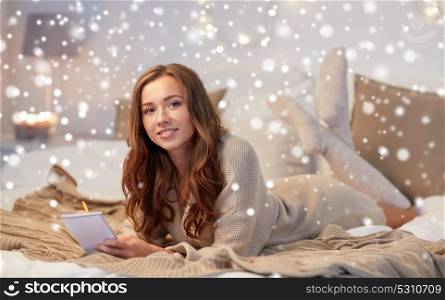 christmas, winter, holidays and people concept - happy young woman with pencil and notebook writing in bed at home bedroom over snow. happy young woman with notebook in bed at home