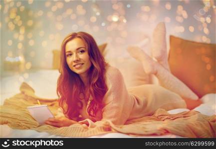 christmas, winter, holidays and people concept - happy young woman with pencil and notebook writing in bed at home bedroom. happy young woman with notebook in bed at home