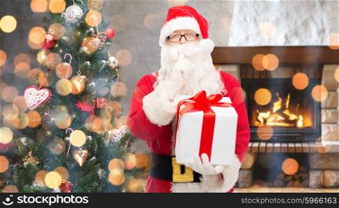 christmas, winter, holidays and people concept - man in costume of santa claus with gift box and tree making hush gesture over home fireplace and lights background