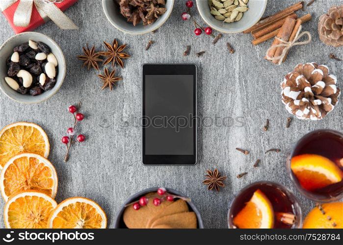 christmas, winter holidays and technology concept - smartphone, hot mulled wine, dry orange slices and aromatic spices on grey background. smartphone, hot mulled wine and christmas spices