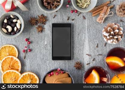 christmas, winter holidays and technology concept - smartphone, hot mulled wine, dry orange slices and aromatic spices on grey background. smartphone, hot mulled wine and christmas spices