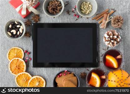 christmas, winter holidays and technology concept - tablet computer, hot mulled wine, dry orange slices and aromatic spices on grey background. tablet computer, mulled wine and christmas stuff