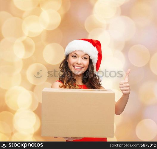 christmas, winter holidays, delivery, gesture and people concept - smiling woman in santa helper hat with parcel box showing thumbs up over beige lights background