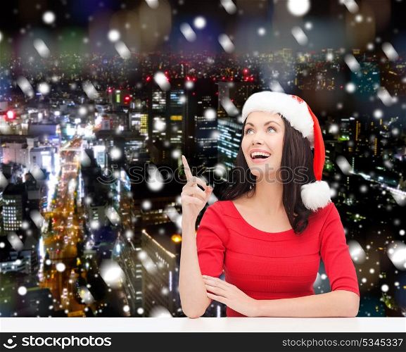 christmas, winter, holidays, happiness and people concept - smiling woman in santa helper hat pointing finger over snowy night city background