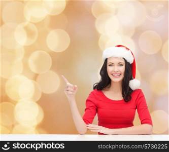 christmas, winter, holidays, happiness and people concept - smiling woman in santa helper hat pointing finger over beige lights background