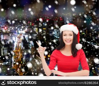 christmas, winter, holidays, happiness and people concept - smiling woman in santa helper hat pointing finger over snowy night city background