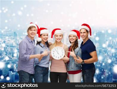 christmas, winter, holidays, time and people concept - group of smiling teenagers in santa helper hats with clock pointing finger over snowy city background