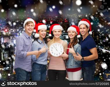 christmas, winter, holidays, time and people concept - group of smiling teenagers in santa helper hats with clock pointing finger over snowy night city background