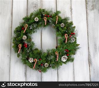 Christmas wreath decorated with pine cones, candy canes, and red berries on rustic white wooden boards.