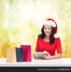 christmas, x-mas, online shopping and electronics concept - smiling woman in santa helper hat with shopping bags and tablet pc computer