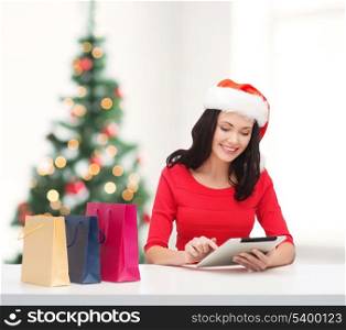 christmas, x-mas, online shopping concept - smiling woman in santa helper hat with shopping bags and tablet pc computer