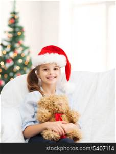 christmas, x-mas, winter, happiness concept - smiling girl in santa helper hat with teddy bear at home