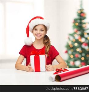 christmas, x-mas, winter, happiness concept - smiling girl in santa helper hat with gift box and wrapping paper