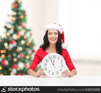 christmas, x-mas, winter, happiness concept - smiling woman in santa helper hat with clock showing 12