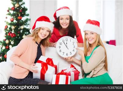 christmas, x-mas, winter, happiness concept - three smiling women in santa helper hats with clock showing 12 and gift boxes