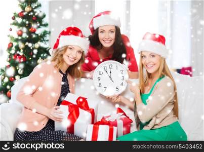 christmas, x-mas, winter, happiness concept - three smiling women in santa helper hats with clock showing 12 and gift boxes