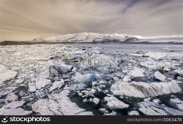 Chunks of ice scattered across a glacier lagoon in Iceland with mountains covered in snow on the background