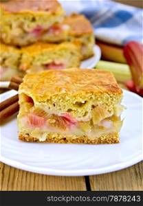 Chunks of sweet cake with rhubarb in a plate, napkin, rhubarb stalks on a wooden boards background