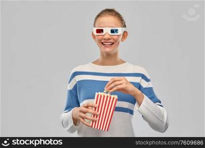 cinema and people concept - happy smiling teenage girl in 3d movie glasses eating popcorn from striped bucket over grey background. teenage girl in 3d movie glasses eating popcorn
